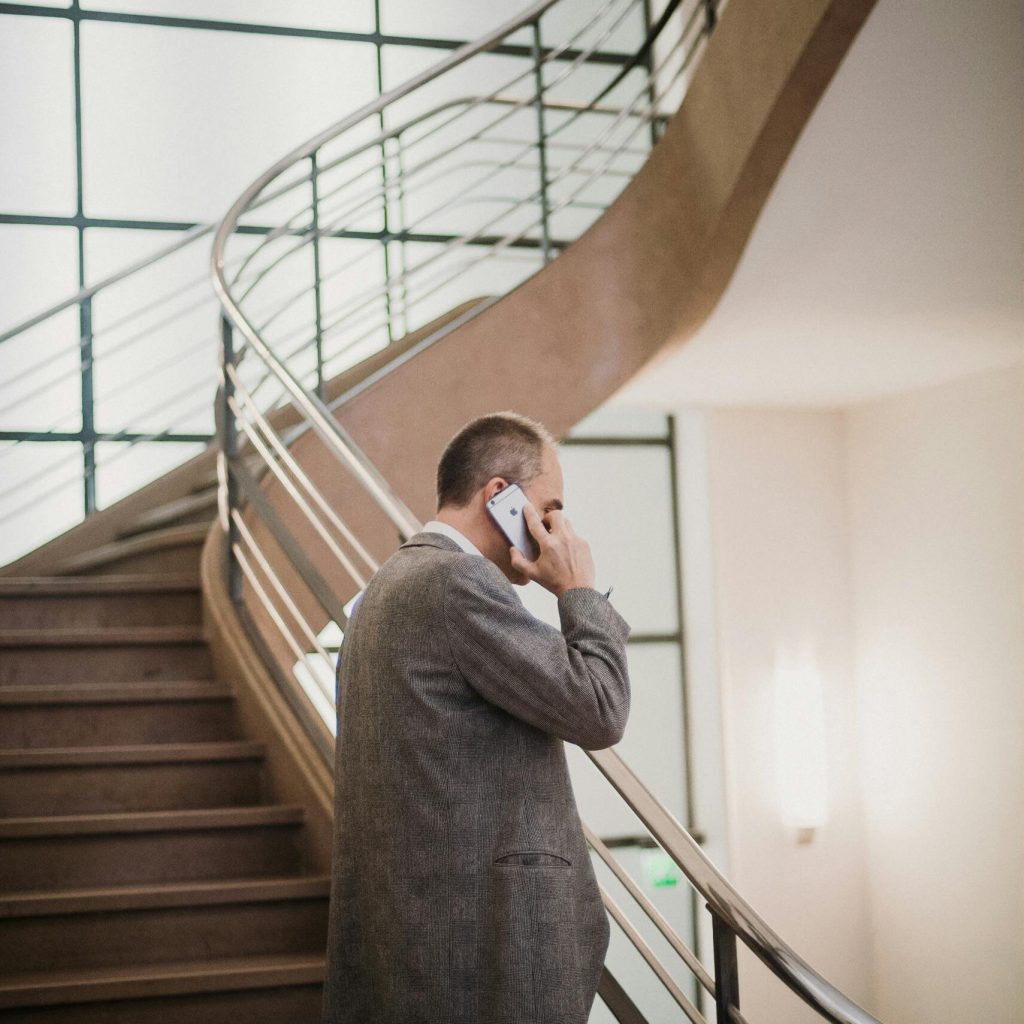 A businessman in a suit talks on his smartphone by a modern staircase indoors.